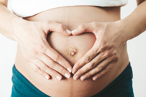 Midsection view of a pregnant woman’s belly. She is doing heart gesture on her belly with her hands.