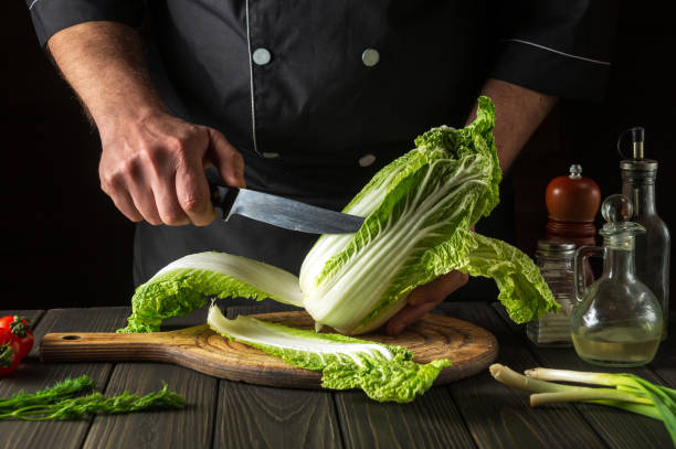 istock Chef or cook cuts fresh napa cabbage with knife for salad on a vintage kitchen table with fresh vegetables. Cooking and restaurant or cafe concept. 1441865781