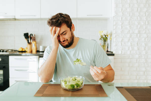 triste jeune homme regarde avec envie la fourchette de salade avec des légumes verts dans la salade à la main devant lui. - longingly photos et images de collection