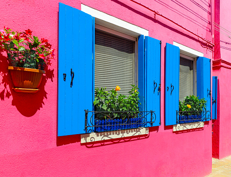 Flower pots decorate on the walls and blue windows of the pink house. Colorful architecture in Burano Island, Venice, Italy
