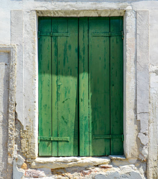 Green window on cracked concrete wall overlooking red bricks in Burano Island, Italy
