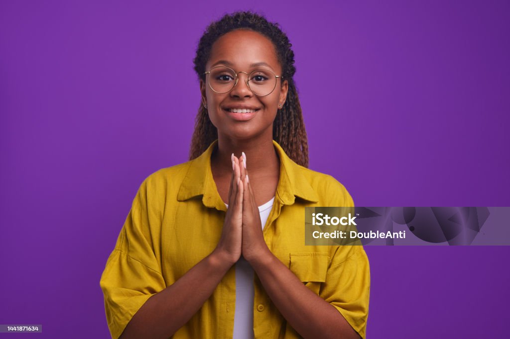 Young sweet good-natured African American woman prayer stands in studio Young sweet good-natured African American woman in casual clothes with smile asks for help doing prayer or wants to thank for support provided to solve problems stands on purple studio background Adult Stock Photo
