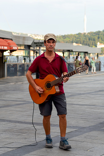 Istanbul, Turkey - August 31, 2022: Male musician Busking playing guitar in Ortakoy district, in front of Bosphorus with Beltas Cafe in the background, and Kucuk Camlica Tower in the far end