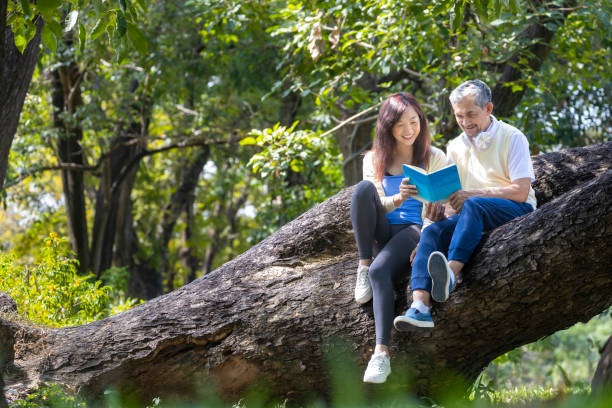 アジアの年配の父と娘が、夏に成熟した木の枝に座って一緒に本を読んで、心身の健康と長寿のコンセプトを主張する - grandparent senior adult child reading ストックフォトと画像
