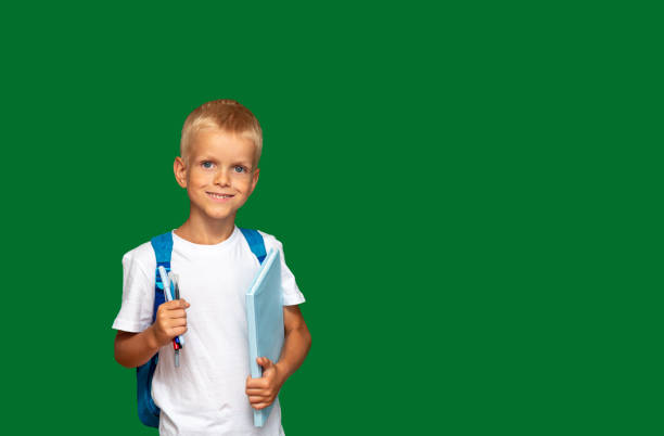 Boy is smiling, with book and markers in his hands, with backpack. Green background with space for text. Selective focus. Picture for articles about children, school, education. stock photo