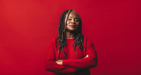 Black woman looking at the camera while standing against a red background with her arms crossed. Mature woman with dreadlocks embracing her natural hair with confidence.