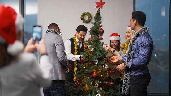 A group of a multiracial business people are posing for photo and video while decorating a Christmas tree at the office after work.