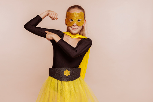 Powerful self confident child girl superhero shows her muscle and strength, isolated over beige background at studio. Wears yellow cape and mask and black outfit. Girl power concept.