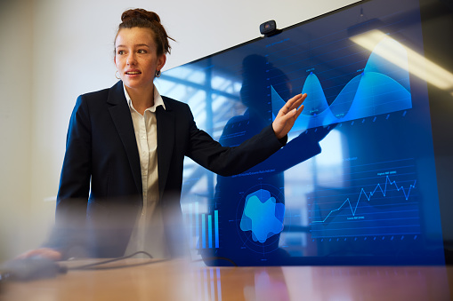 Attractive young female adult is going over  graphs shown on a large computer screen at a business meeting. Waist up close-up, focus on background