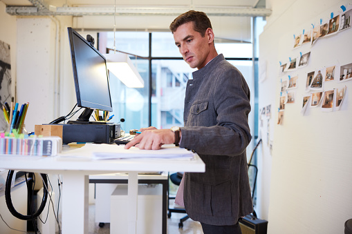 Handsome mid adult male in a modern office working at a standing desk in front of a board with pinned photos. Three quarter length side view close up with copy space