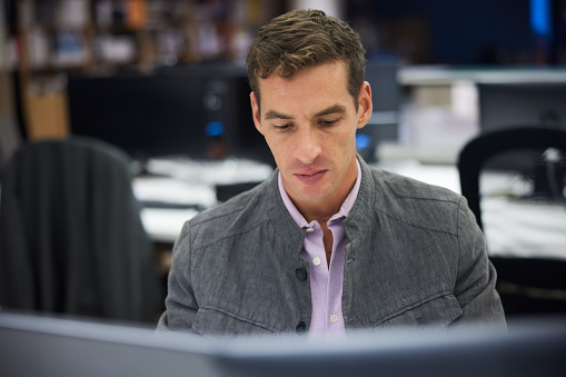 Mid adult male with short dark hair is seen behind the edge of a computer screen seated at desk and looking downward. Focus on foreground with copy space