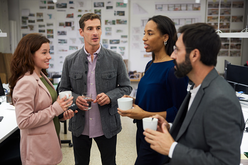 Diverse team members, young adult female, mid adult male, African-American mid adult female and mature male with beard, are standing around with coffee in hand in conversation. Close-up with focus on foreground