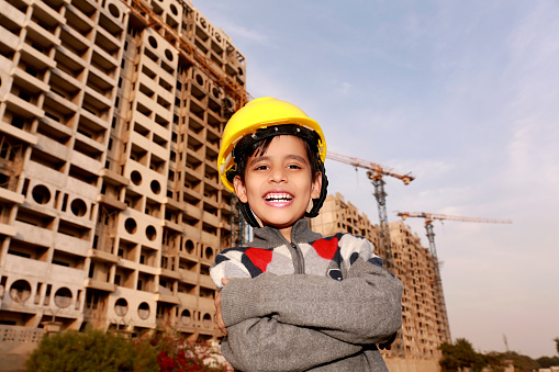 Cute cheerful elementary age Engineer of Indian ethnicity standing with arm crossed wearing safety helmet portrait near construction site with arm crossed.