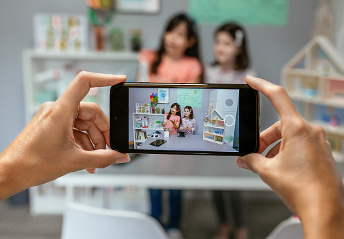 Hands of unrecognizable teacher recording video in mobile phone of female students showing led lamp in ecology classroom. Selective focus on mobile screen.