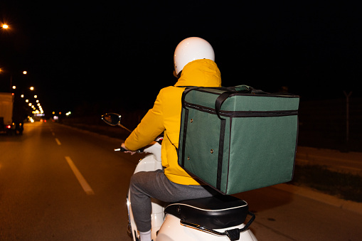 Rear-view of unrecognizable Caucasian man, an delivery person, driving scooter, while delivering the orders around the city