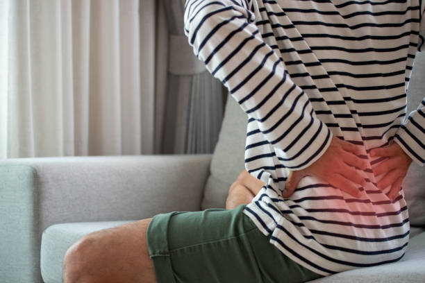 working man sitting on the sofa in the living room at home she used both hands to press down on the lower back. she is suffering from back pain from sitting for a long time. - lower back pain imagens e fotografias de stock