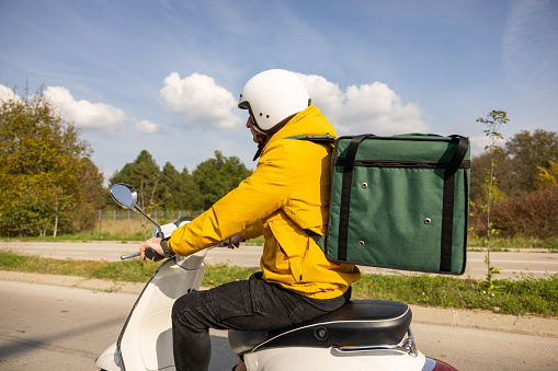 Side view of Caucasian man, an delivery person, driving scooter, while delivering the orders around the city