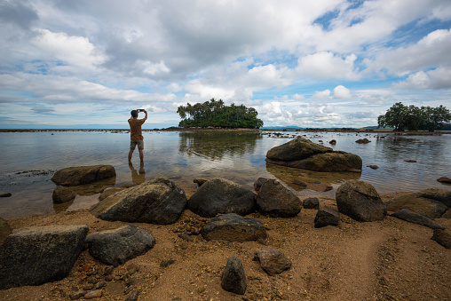 A senior Asian male tourist taking photograph at small island on Naiyang beach, Phuket, Thailand.