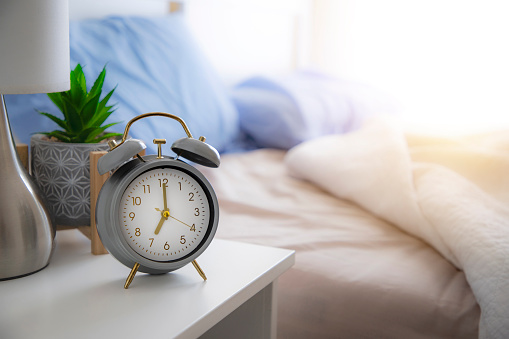 Front view of an alarm clock on a nightstand with an unmade and defocused bed background in the morning