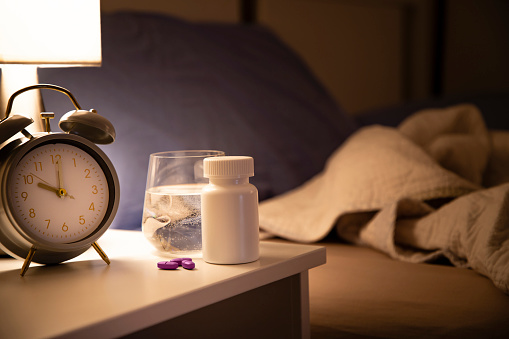 Front view of a pill container and an alarm clock on a nightstand with an unmade and defocused bed background at night