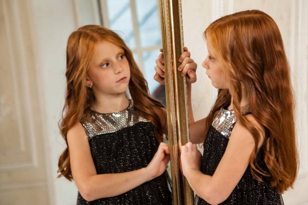 bambina carina di 5-6 anni con capelli rossi, in posa, guardando riflesso, modello di moda in elegante vestito nero elegante in salotto allo specchio. giovane attrice alla moda a casa. copiare lo spazio di testo - 6 7 years little girls child portrait foto e immagini stock