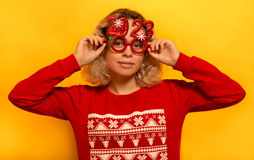 Young blonde woman wearing red Christmas sweater with ornament pattern, holding party glasses and looking at camera on a yellow background.

Happy holiday and New Year 2023 celebration concept.