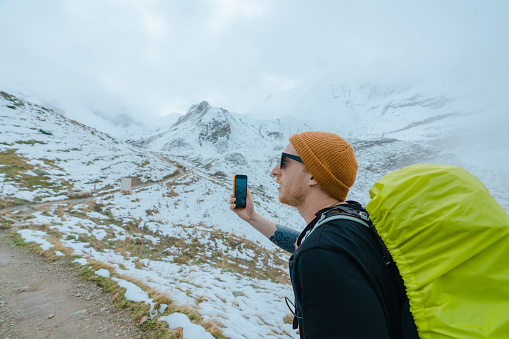 Young Caucasian man photographing with smartphone while hiking in snow-covered mountains