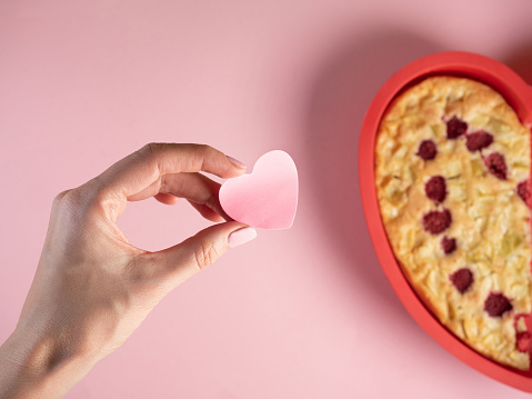 Woman hand hold heart shape pink paper, heart shape half apple pie on pink background. Celebrating Valentine's Day