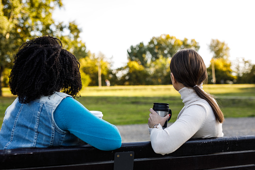 Rear view of two happy young diverse women, friends, sitting on the street bench, having coffee from reusable mugs, bonding, chatting and enjoying a sunny day outside.