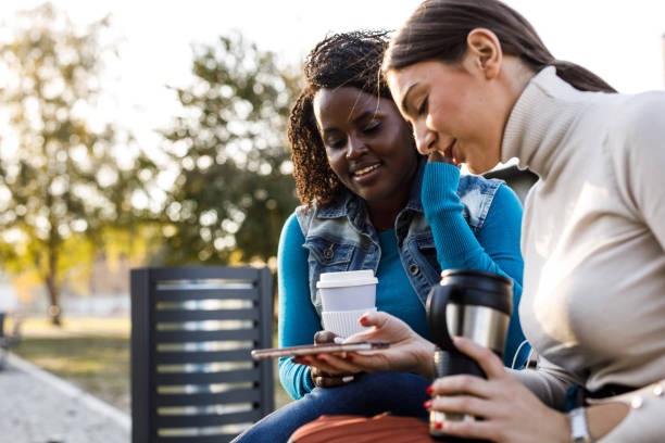 istock Two diverse friends sitting on a street bench, having coffee and looking at something on smart phone 1441908764