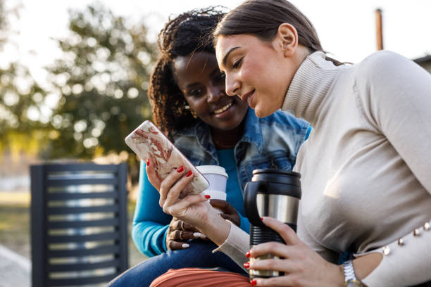 istock Two diverse friends sitting on a street bench, having coffee and looking at something on smart phone 1441908840