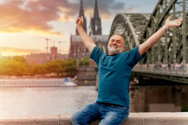a pensioner traveling across europe by train. bearded man  sitting on embankment of Rhine on background of Cologne Cathedral and Hohenzollern Bridge in Koel, Germany. Tourism and travel by Germany concept