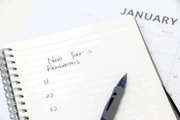 Photo of Writing new years resolutions, list on notepad with ballpoint pen, January calendar in background, selective focus. Setting goals for the new year.