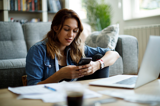 Young smiling woman using mobile phone while studying at home