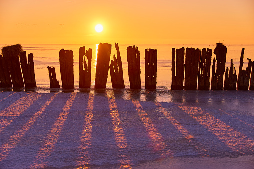 Old wooden breakwater in frozen lake during sunset, Netherlands. The sky is clear and there is some snow over the ice. The sunlight casts shadows on the ice.