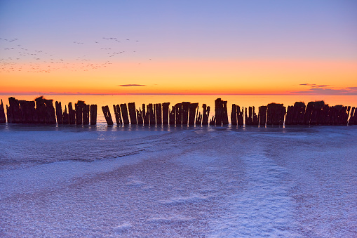 Old wooden breakwater in frozen lake during sunset, Netherlands. The sky is clear and there is some snow over the ice.