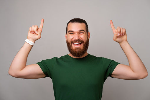 Cheerful bearded man wearing green T shirt is pointing up above his head on grey background.