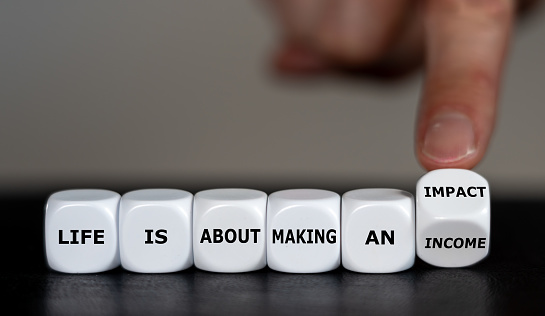 Hand turns dice and changes the expression 'life is about making an income' to 'life is about making an impact'.