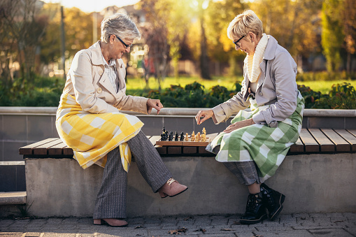 Two senior adult women playing chess on the bench outdoors in the park.