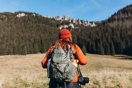 Back view of stylish woman wearing green backpack and red hat looking at mountain view while relaxing in nature. Travel and wanderlust concept