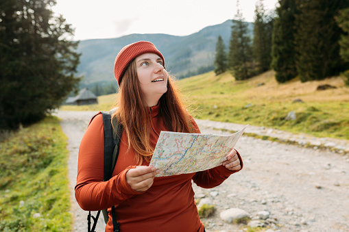 Stylish woman holding paper map relaxing in nature. Travel and active lifestyle concept. Hiking active female lifestyle wearing backpack and knit hat exercising outdoors.