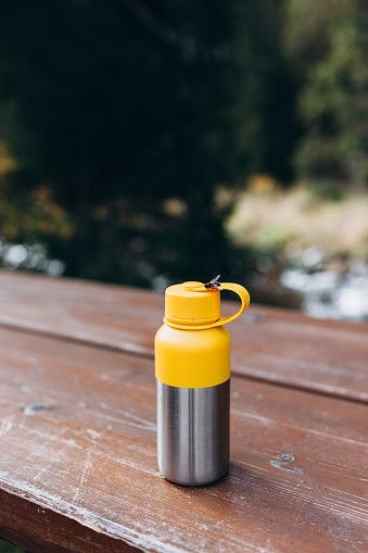 A vacuum thermos stands on a tree desk in the wild. Hiking equipment on footpath in woodland. Travel outdoor concept