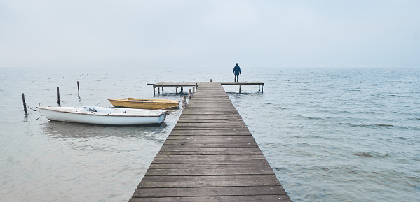 Man standing on a wooden jetty or pier at a lake at foggy morning.
