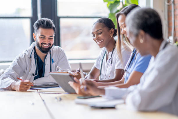 Medical Team Meeting A small group of four medical professionals sit around a boardroom table as they meet to discuss patient cases.  They are each dressed professionally in scrubs and lab coats as they focus on working together. physician computer stock pictures, royalty-free photos & images