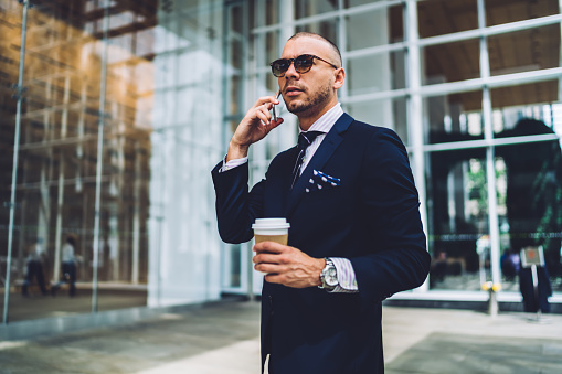 Serious smart businessman in elegant suit and sunglasses with coffee on go talking on mobile phone in light glass business hall