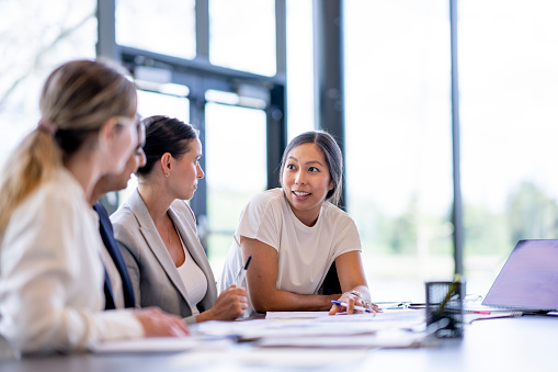 A small group of four female business professionals sit together at a boardroom table as they meet to discuss and strategize.  They are each dressed professionally in business attire and are focused on their objective.