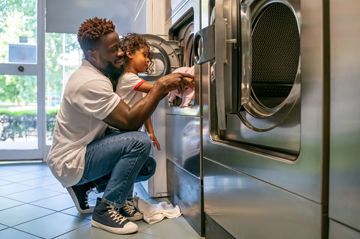 Focused little girl putting dirty clothes into the automatic front-load washing machine assisted by her pleased parent