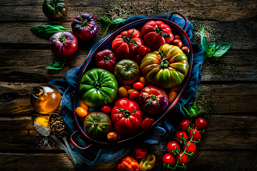Vegetables: overhead view of different kind of organic tomatoes in a tray shot on rustic wooden table. Olive oil, peppercorns, salt and herbs complete the composition. High resolution 42Mp studio digital capture taken with SONY A7rII and Zeiss Batis 40mm F2.0 CF lens