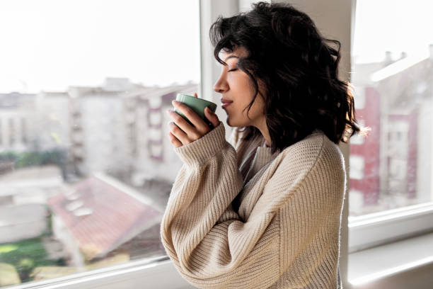 istock Young woman enjoying cup of coffee while looking through  window at home 1441995638