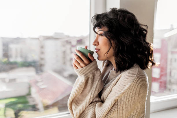 istock Young woman drinking cup of coffee while looking through window 1441996206